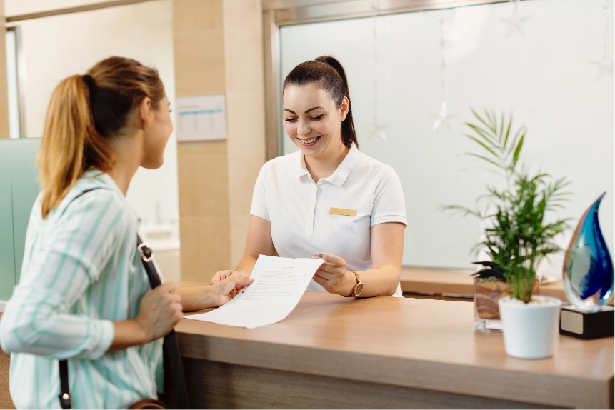 Smiling receptionist handing paperwork to a female patient at dental clinic front desk