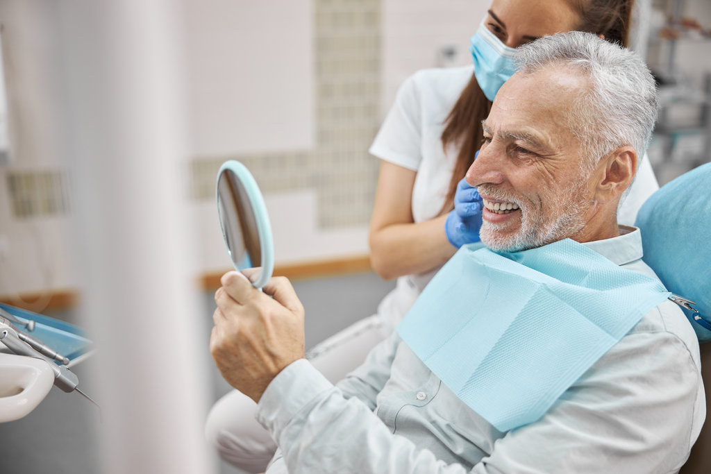 Joyous elderly man sitting in a dental chair with a doctor by his side while looking in the mirror