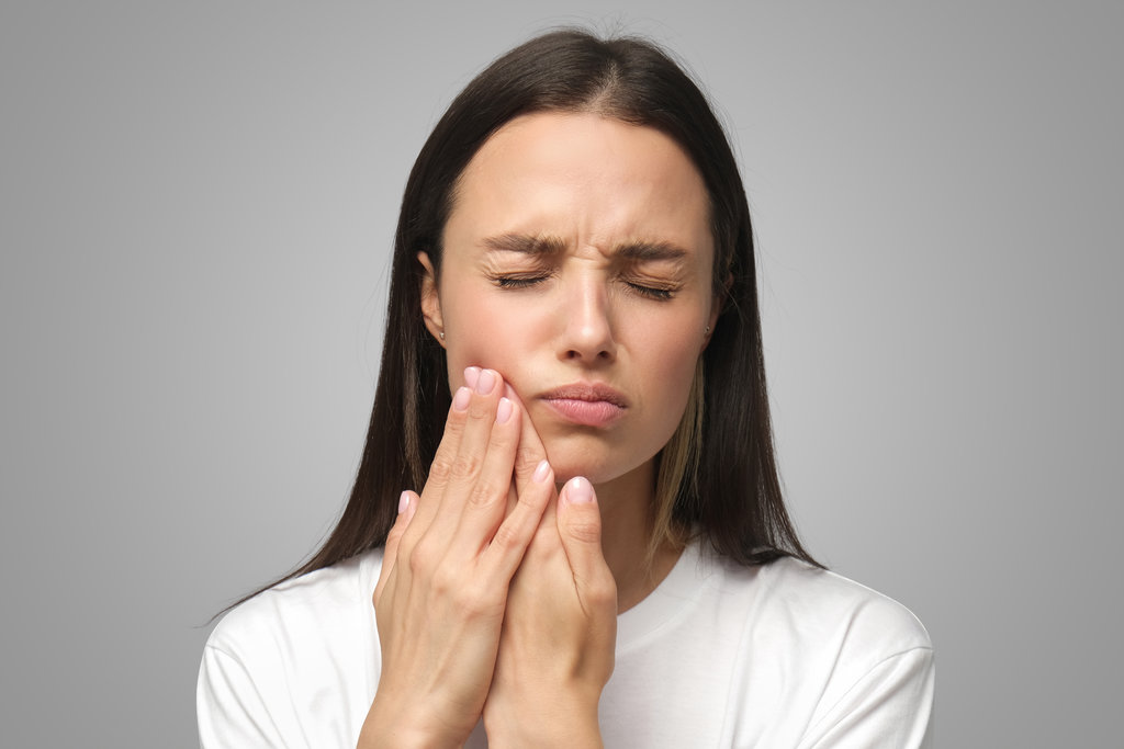Young miserable woman experiencing severe toothache, pressing palm to cheek, closing eyes because of strong pain, isolated on gray background
